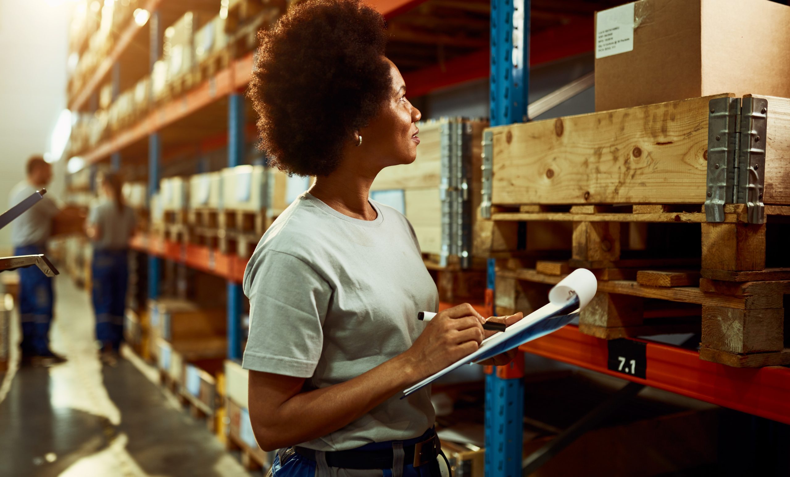 Black female warehouse worker taking notes while checking stock in a warehouse.