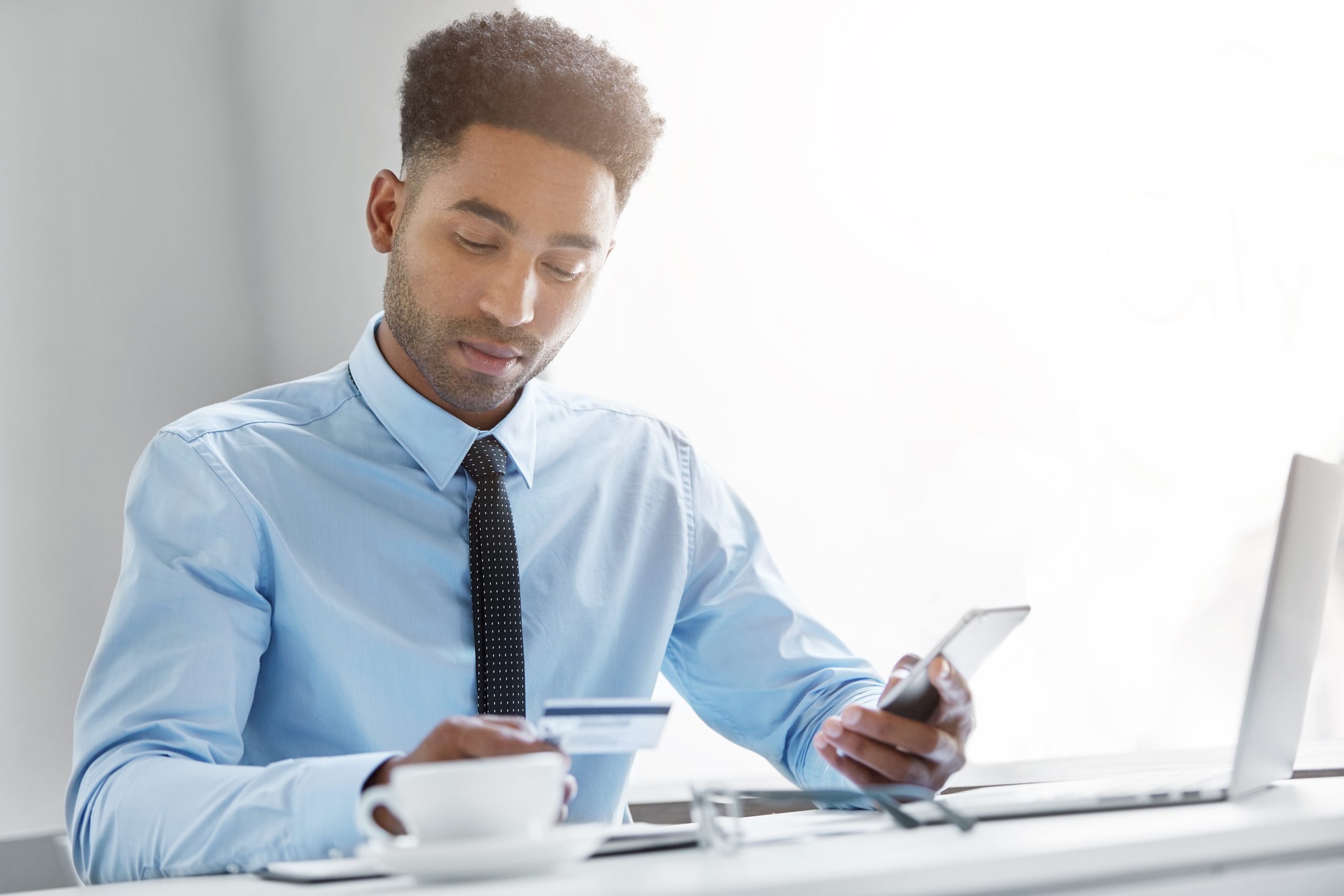 Picture of attractive young African American manager dressed in formal wear sitting at his workspace with cell phone, looking at information on his credit card while signing up for mobile banking app